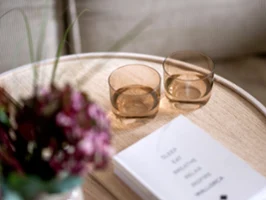 Like Glass Clay tumblers on a wooden coffee table Two minimalist, rounded Like Glass crystal tumblers in the warm amber shade Clay by Villeroy & Boch, placed on a light wooden side table next to a white book and a blurry bouquet of bordeaux-colored flowers.