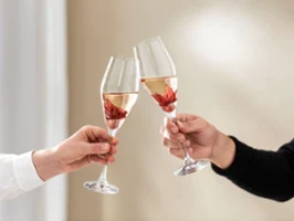 Against a neutral background, two people toast with a glass of La Divina from Villeroy & Boch, each holding a hibiscus flower.
