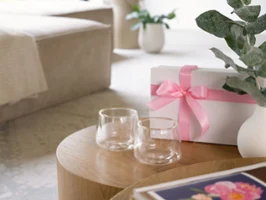 Elegant Hot & Cold Beverage Glasses with Gift Box Two double-walled glasses with gold rims placed on a wooden table, next to a white gift box with a pink ribbon and a white vase with green eucalyptus leaves in a modern, cozy living room.
