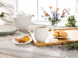 Eine Teetasse von Villeroy & Boch New Cottage auf einem Tablett mit Keksen Eine Teetasse von Villeroy & Boch New Cottage auf einem Tablett mit Keksen, Teekanne, Kandiszucker und getrockneten Orangenscheiben auf einem mit Blumen geschmückten Holztisch.
