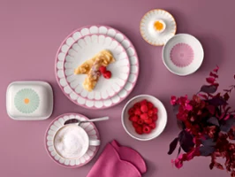 Overhead flat lay of Villeroy & Boch's Fleur pink porcelain plates with a crepe, a cup of cappuccino, a butter dish, and small bowls filled with fresh raspberries and a soft-boiled egg, set on a dusty pink background next to magenta foliage.