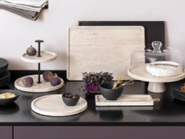 Küchenarbeitsplatte mit eleganten Manfacture Travertin Tabletts, auf denen Feigen, Brie, Brot und eine Schüssel Gemüse präsentiert werden. 