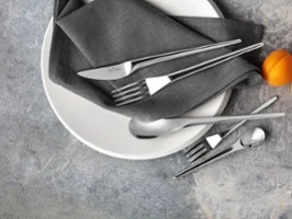 Ein weißer NewMoon Teller mit einer grauen Serviette, NewMoon Besteck und einer Aprikosen auf einer grauen, strukturierten Oberfläche.