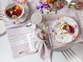 Mariefleur auf einem Tisch Ein mit Blumenmuster gedeckter Tisch mit Tellern, Besteck, einer Serviette, einem kleinen Dessert und einer Obstschale auf einer hellen Tischdecke.