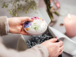 Hands are holding a Mariefleur bowl by Villeroy & Boch with a floral pattern, being unwrapped next to a burning pink candle.