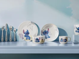 Une série d'assiettes et de tasses de la collection Disney Stitch sont décorées de motifs de dessins animés et exposées sur une étagère bleu clair avec des ustensiles.