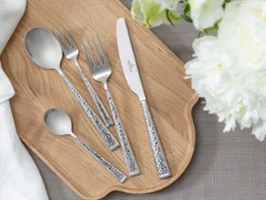 Draufsicht auf Blacksmith Besteck – Esslöffel, Tafelgabel, Tafelmesser, Kaffeelöffel und Dessertgabel – ordentlich auf einem Holzbrett arrangiert. Umgeben von feiner weißer Blumendeko. Eine saubere, natürliche und elegante Ästhetik.