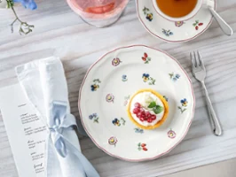 Petite Fleur Teller mit einer Torte, einer Teetasse, einem Getränk mit einer Blume und einer Serviette auf einem Holztisch.