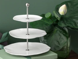 Three-tiered white Pura étagère with metal handle next to a lotus plant Three-tiered white Pura étagère with metal handle, next to it a large green lotus plant and a closed gift box.