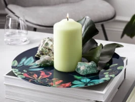 A still life scene with a lit candle, crystals and foliage on a decorative Avarua plate, with books and a pen on a modern table.