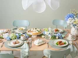 Une table à manger pour quatre personnes décorée dans des tons pastel, avec des assiettes remplies de plats, des tasses à thé, des compositions florales bleues et blanches et une vaisselle élégante, créant une atmosphère fraîche et accueillante.
