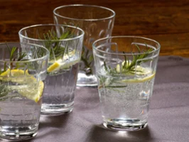 Vasos llenos de agua Cuatro copas "Dressed Up" de Villeroy & Boch llenas de agua con gas, adornadas con rodajas de limón y ramitas de romero, colocadas sobre un paño gris.