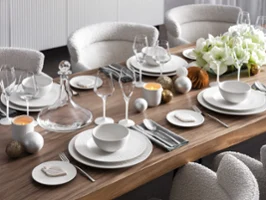 Una mesa de comedor moderna está festivamente puesta con platos blancos Manufacture Rock Blanc, vasos, servilletas grises, velas y una corona de flores blanca. 