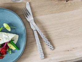 On a light-coloured wooden table, there is a Lave plate with salad next to a Boston fork and a Boston knife.