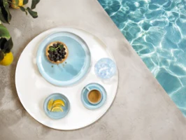 Okrągła taca z naczyniami z kolekcji Perlemor Aqua i szklanką wody, na brzegu basenu.
