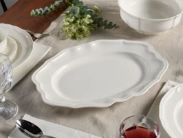 Manoir Oval Platter A white platter with scalloped edges is elegantly placed on a set dining table adorned with a tablecloth, surrounded by plates, glasses, cutlery, and bowls.