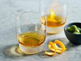 The Octavie Tumblers Gift Set, featuring two elegant crystal glasses filled with whisky, is complemented with a Manufacture Rock bowl filled with green olives.