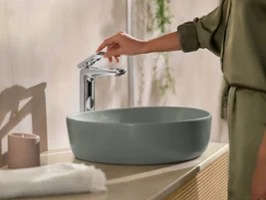 Une personne touche une robinetterie en chrome, installée sur un lavabo ovale vert sur pied.