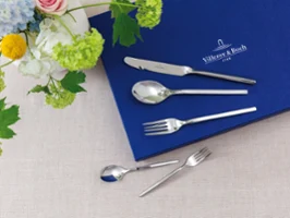 Besteckset mit Messer, Löffel und Gabel auf blauem Tuch neben Blumenarrangement auf beigem Untergrund.	