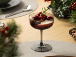 Primer plano de una copa de champán Like Glass de Villeroy & Boch en color Smoke, utilizada como copa de postre para una crema navideña en capas con cerezas rojas y una ramita de romero, sobre una mesa de comedor moderna.