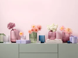 A series of pastel-coloured vases from the Fleur collection in various shapes and sizes with pink, white and peach-coloured flowers, standing on a shelf with stacked books and small boxes.
