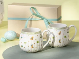 Dos tazas Easter Delight con motivos florales colocadas delante de una caja de regalo atada con una cinta azul claro.