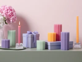A collection of colorful decorative candles from the Fleur collection, in various shapes and pastel shades, stands on a green surface with pink flowers in the background in front of a light pink wall.