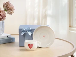Tasse blanche With Love avec motif cœur rouge, soucoupe assortie et boîte cadeau bleue avec nœud sur une table en bois près de la fenêtre.