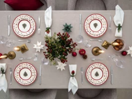 A dining table setting for four people with festive decorations, including Toys Delight plates with Christmas tree patterns, red and gold accents, folded white napkins, candles and a centrepiece with red berries and green plants.