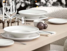 A dining table setting with white Afina plates, bowls and glasses next to a Christmas tree decorated with lights and ornaments.