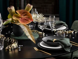 Elegant Villeroy & Boch table setting with dark Manufacture plates, gold cutlery, green napkins and a black Collier vase with tropical flowers and leaves. Gold streamers and glasses decorate the black table.
