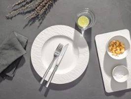 Ein weißer Teller mit Gabel und Messer, ein Glas Wasser mit Zitrone sowie ein Tablett mit Müsli und einer Schüssel auf einem grauen Untergrund.