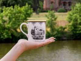 One hand holds a decorative Audun cup with an illustration of a building in front of a river and trees.