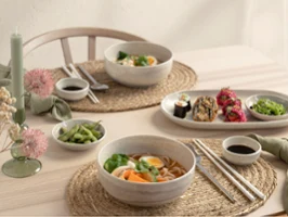 A table set for two with bowls of ramen, sushi rolls, edamame, chopsticks and dipping sauces from the Perlemor collection on woven placemats.