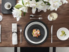 Vista cenital de una mesa contemporánea que combina la colección Manufacture Rock en negro mate—con un plato gourmet, una bandeja rectangular y un plato de desayuno—con la línea minimalista NewMoon en blanco brillante, incluyendo plato llano, plato de pan y cuenco para salsas. Las formas y superficies contrastadas crean una composición artesanal llamativa y armoniosa.