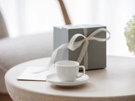 Filiżanka do espresso ze spodkiem Afina w pudełku upominkowym Biała filiżanka Afina na spodku, pudełko prezentowe z kokardą i bukiet białych kwiatów na okrągłym stole.