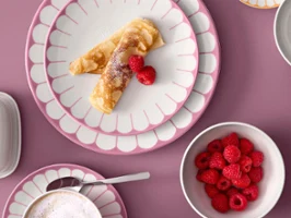 Vogelperspektive eines stilvollen Frühstückstischs mit der Fleur-Kollektion von Villeroy & Boch in Pastellrosa, Minzgrün und Orange. Zu sehen sind Teller mit Crêpes und Himbeeren, eine Cappuccino-Tasse, eine Butterdose, ein Eierbecher und eine Schale mit frischen Himbeeren auf einem altrosa Hintergrund.