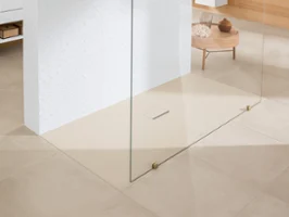 Une salle de bains minimaliste avec des carreaux de sol beiges, une douche à l’italienne séparée par une plaque en verre, un mur carrelé blanc et une petite table en bois avec des objets décoratifs en arrière-plan.