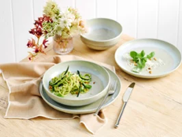 The Perlemor Alga dinner set for one, featuring a plate filled with fresh zucchini pasta, rests on a brown linen tablecloth atop a rustic wooden table, accompanied with silver cutlery.