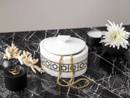 A decorative, gold-patterned MectroChic sugar bowl on a marble surface, surrounded by dried coral, a candle, a flower and a ring on a black lid.