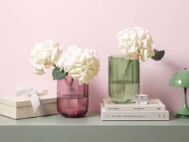 Two Fleur glass vases with white hydrangeas on a table next to stacked books, a gift box and a small green lamp.
