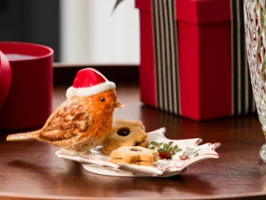On a festively designed Villeroy & Boch plate are a bird figurine with a Christmas hat and some cookies, next to it are a wrapped gift and a glass vase.