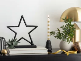 Sobre una mesa con objetos decorativos hay una escultura de una estrella negra, una vela a rayas, una plantita y una lámpara dorada.