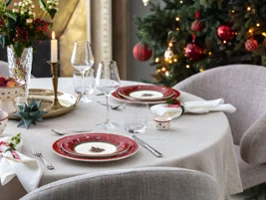 Una mesa de comedor redonda preparada para una comida navideña con la vajilla Toy's Delight, una vela encendida y un árbol de Navidad decorado al fondo.