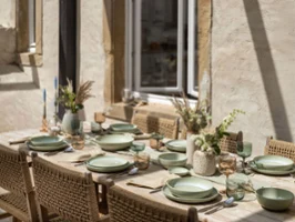 A summery dining table with green plates from the Perlemor collection. A summery dining table with green plates from the Perlemor collection, glasses and cutlery on a wooden surface. It is framed by wicker chairs and the centrepiece is adorned with decorative vases full of flowers.