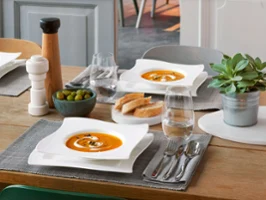A dining table set with plates from the NewWave collection by Villeroy & Boch with tomato soup, slices of bread, olives, cutlery, two glasses of water, a pepper mill and a potted plant.