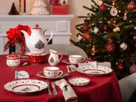 A table set with crockery and cutlery featuring Christmas motifs, next to a decorated Christmas tree and an illuminated mantelpiece.