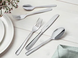 Liam flatware on a white wooden table A neatly arranged stainless steel flatware set with knives, forks, and spoons on a light-colored wooden surface. Next to it are a white plate and a green napkin, with a flower arrangement in the top left-hand corner.