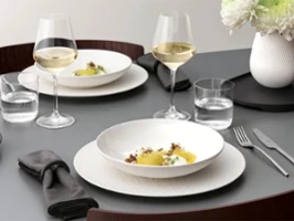 A formal table setting with a white bowl on a white plate, glassware, and silver cutlery on a grey table and food.