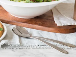 Daily Line Salatbesteck A metal spoon and a metal fork are lying on a cloth napkin next to a salad bowl and a plate on a marble table.